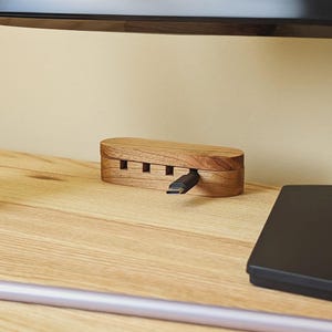 May include: A wooden USB hub with a natural finish. The hub has a rectangular shape with rounded edges and four slots on the front. A black USB-C cable is plugged into one of the slots. The hub is on a wooden desk.