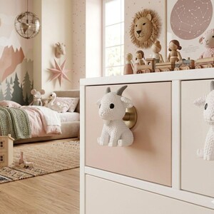May include: A white chest of drawers with goat-shaped handles. The drawers are painted in light pink and white. The room has a children's theme, with a bed, toys, and wall art.