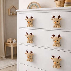 May include: A white dresser with six knitted reindeer drawer pulls. Each reindeer has brown antlers, a black bead eye, and an orange nose. The reindeer are beige and are attached to the white drawers. The dresser is in a nursery setting.