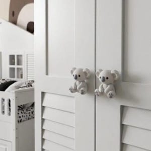 May include: A white wooden wardrobe featuring two louvered doors with two light gray koala-shaped knobs. Each koala figure is seated with visible paws and ears, serving as functional handles for the cabinet doors on this piece of furniture.