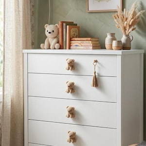 May include: A white five-drawer dresser featuring light brown crocheted bear-shaped knobs on the left side of each drawer and a single gold-toned metal ring pull with a matching beige tassel on the top right drawer.