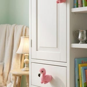 May include: A white cabinet with pink flamingo-shaped drawer pulls. The cabinet has a door and drawers. The flamingo pulls are made of a textured material and have black beaks. Bookshelves are visible to the right.
