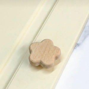 Könnte beinhalten: Ein hölzerner Schrankknauf in Blumenform. Der hellbraune Knauf hat ein fünfblättriges Design. Er ist an einer cremefarbenen Schranktür befestigt. Der Knauf ist ein dekoratives Element für Möbel.