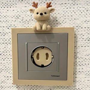 May include: A beige crochet deer figure sits atop a square electrical wall outlet cover. The outlet features a grey faceplate with the brand name 'netelsan' printed in black, and a circular cream-colored socket insert with two vertical slots.