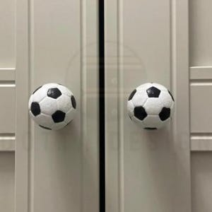 May include: Two white and black football-shaped cabinet knobs are mounted on a white cabinet door. The knobs have a classic football design with black pentagons and white panels. The cabinet doors are closed.