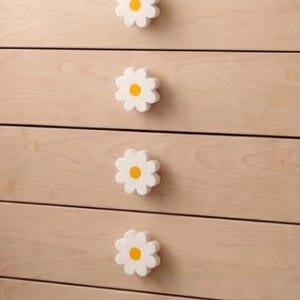 Könnte beinhalten: Eine Kommode aus hellem Holz mit fünf Schubladen, die jeweils mit einem weißen, blumenförmigen Knopf mit gelber Mitte versehen sind. Die Kommode hat eine glatte Oberfläche mit natürlicher Holzmaserung und ein klares, horizontales Design.