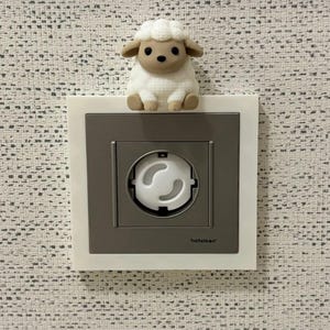 May include: A white and beige sheep figurine sits atop a square electrical wall outlet. The outlet features a gray faceplate with a white frame and a white child safety plug cover inserted into the center. The brand name netelsan is printed.