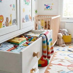 May include: A white chest of drawers with open drawers containing folded children's clothes and toys. The drawers are decorated with animal stickers. A cot and colourful rug are in the background, creating a nursery setting.