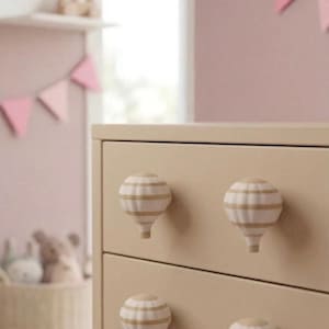 May include: A beige chest of drawers with four drawers, each fitted with a hot air balloon-shaped knob. The knobs are white with beige stripes. The background includes a pink wall, a shelf, and a pink triangle garland.