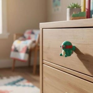 May include: A green turtle-shaped drawer knob with a red eye mask, mounted on a light-toned wooden dresser drawer. The knob features a smooth, rounded design with a cream-coloured belly and small limbs, providing a functional handle for furniture.