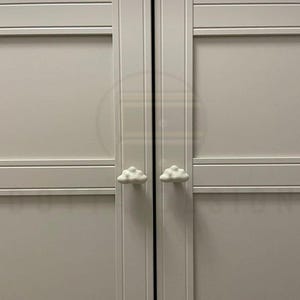 May include: Two white cloud-shaped cabinet knobs on a light grey cabinet door. The knobs are positioned centrally on the door panels, adding a playful element to the furniture. The cabinet doors feature horizontal paneling.