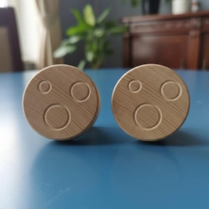 May include: Two round, wooden cabinet knobs with a light brown finish. Each knob features a carved design of three concentric circles. The knobs are displayed on a blue surface, suggesting a home decor or hardware theme.