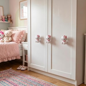 Könnte beinhalten: Ein weißer Kleiderschrank mit drei Schrankknöpfen in Form der Hello Kitty-Figur, jeweils mit weißem Körper, rosa Schleife und schwarzen Gesichtszügen, die einen dekorativen Akzent an dem glatten, einfarbig weißen Möbelstück mit Doppeltür setzen.