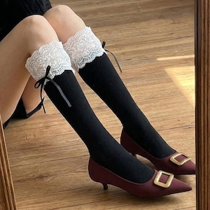 Pu&ograve; includere: Calzini neri al ginocchio con finiture in pizzo bianco e fiocchi neri. I calzini sono abbinati a tacchi bordeaux a punta con fibbie quadrate dorate. Le scarpe hanno un tacco basso. L'immagine si riflette in uno specchio.