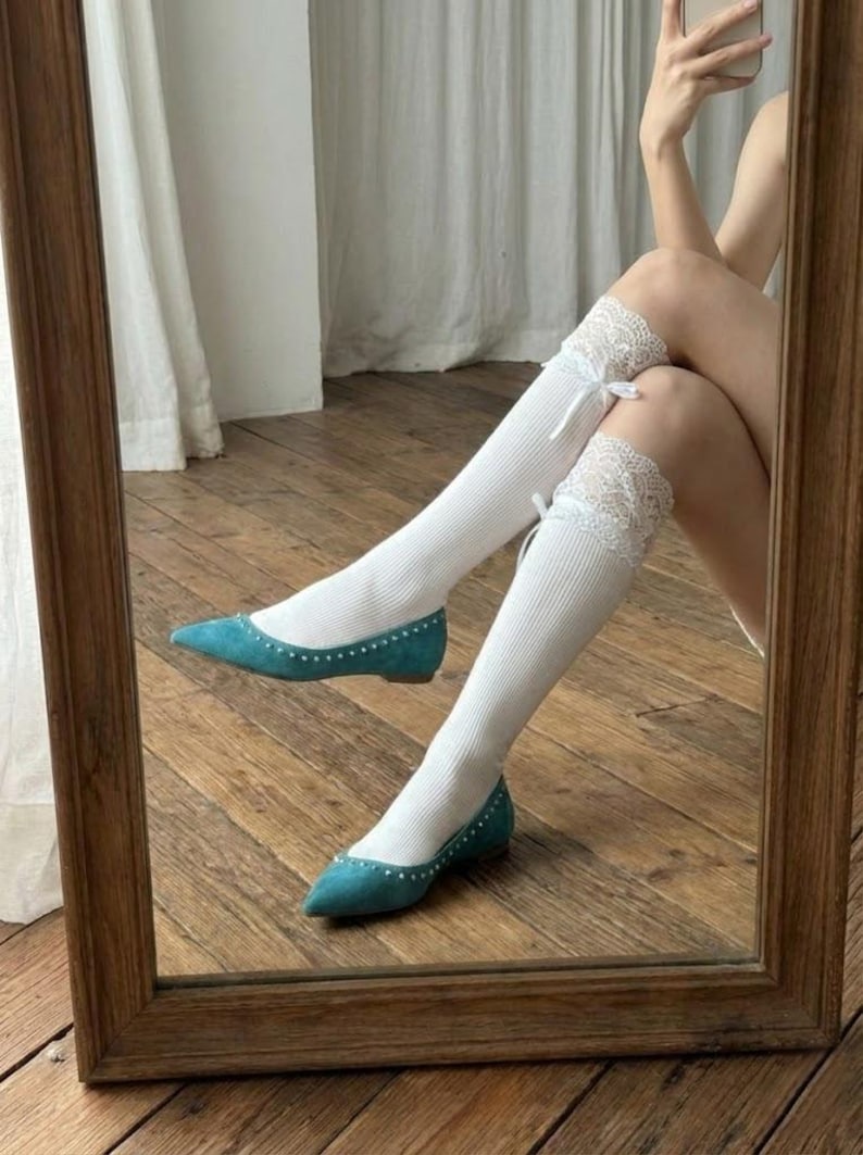 Pu&ograve; includere: Uno specchio riflette ballerine turchesi con borchie e calze al ginocchio bianche. Le calze hanno un bordo in pizzo e un piccolo fiocco. Le ballerine hanno la punta a punta.