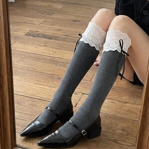 Pu&ograve; includere: Scarpe Mary Jane nere in vernice con tacco basso, abbinate a calze al ginocchio grigie. Le calze sono rifinite con pizzo bianco e nastri neri. L'immagine &egrave; riflessa in uno specchio con cornice in legno.