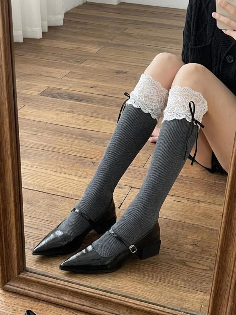 Pu&ograve; includere: Scarpe Mary Jane nere in vernice con tacco basso, abbinate a calze al ginocchio grigie. Le calze sono rifinite con pizzo bianco e nastri neri. L'immagine &egrave; riflessa in uno specchio con cornice in legno.