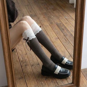 Pu&ograve; includere: Calze al ginocchio marroni con finiture in pizzo bianco e un piccolo fiocco marrone. Le calze sono abbinate a mocassini neri con cinturino bianco. L'immagine si riflette in uno specchio con cornice in legno.