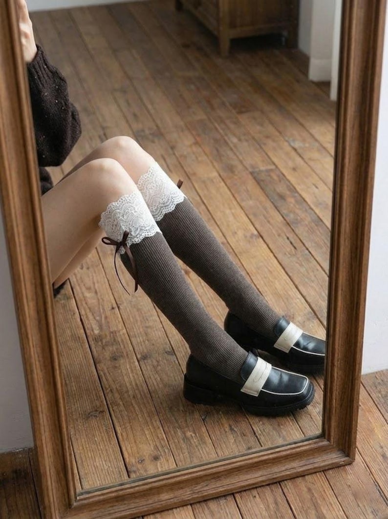 Pu&ograve; includere: Calze al ginocchio marroni con finiture in pizzo bianco e un piccolo fiocco marrone. Le calze sono abbinate a mocassini neri con cinturino bianco. L'immagine si riflette in uno specchio con cornice in legno.