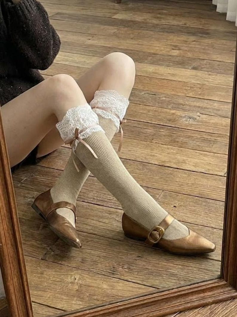 Pu&ograve; includere: Calze al ginocchio color beige con bordi in pizzo bianco e piccoli fiocchi rosa. Sono abbinate a scarpe Mary Jane color bronzo con fibbia. Scarpe e calze su una superficie di legno.