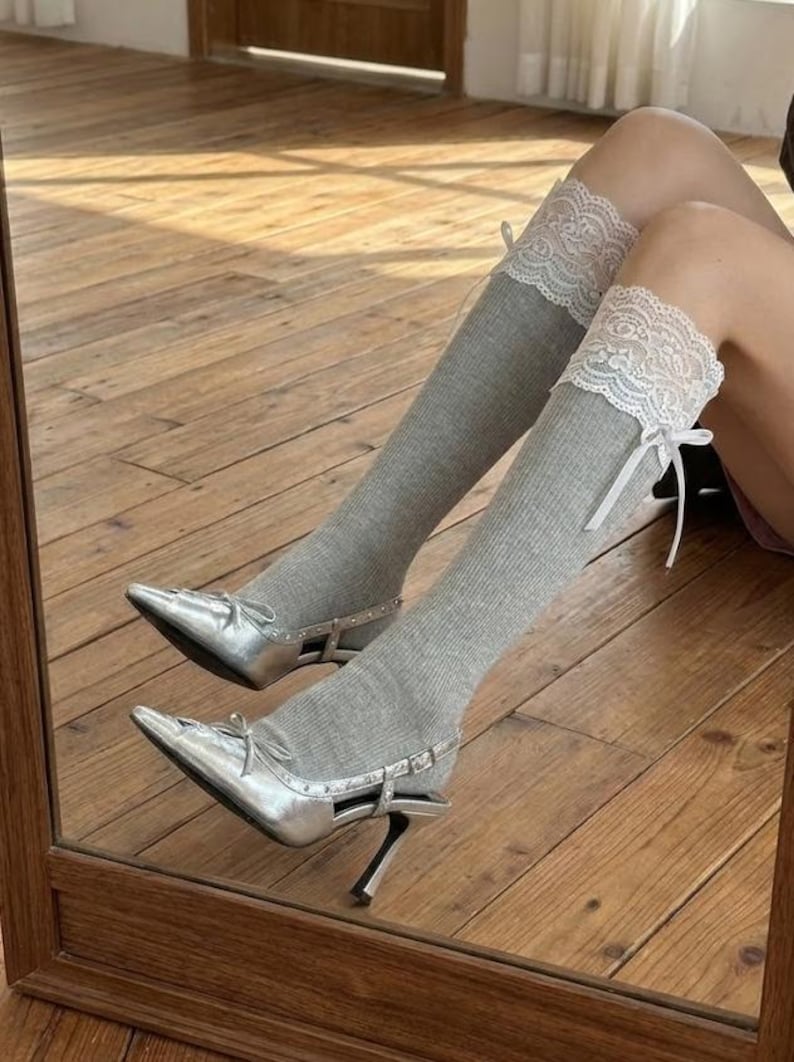 Pu&ograve; includere: D&eacute;collet&eacute; argentate a punta con fiocchi e calze al ginocchio grigie a coste. Le calze hanno finiture in pizzo bianco e nastri bianchi. Le scarpe si riflettono in uno specchio con cornice in legno.