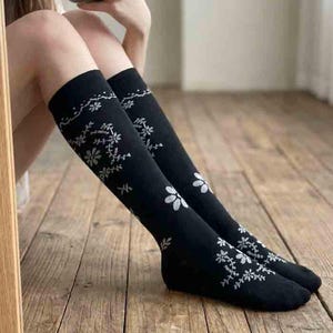 May include: Black knee-high socks with white floral patterns. The socks feature a repeating design of flowers and vines along the top and down the front. The socks are made of a soft material and are suitable for casual wear.