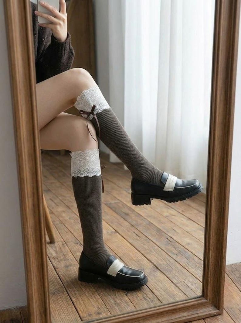 Pu&ograve; includere: Calze al ginocchio grigio scuro con finiture in pizzo bianco e un piccolo fiocco marrone. Le calze sono abbinate a mocassini neri con cinturino bianco. L'immagine &egrave; riflessa in uno specchio con cornice in legno.