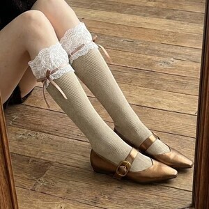 Pu&ograve; includere: Calzini al ginocchio a coste beige con finiture in pizzo bianco e piccoli fiocchi marroni. I calzini sono abbinati a scarpe Mary Jane color bronzo con chiusure con fibbia. L'immagine &egrave; ripresa da una prospettiva bassa, mostrando gambe e piedi.