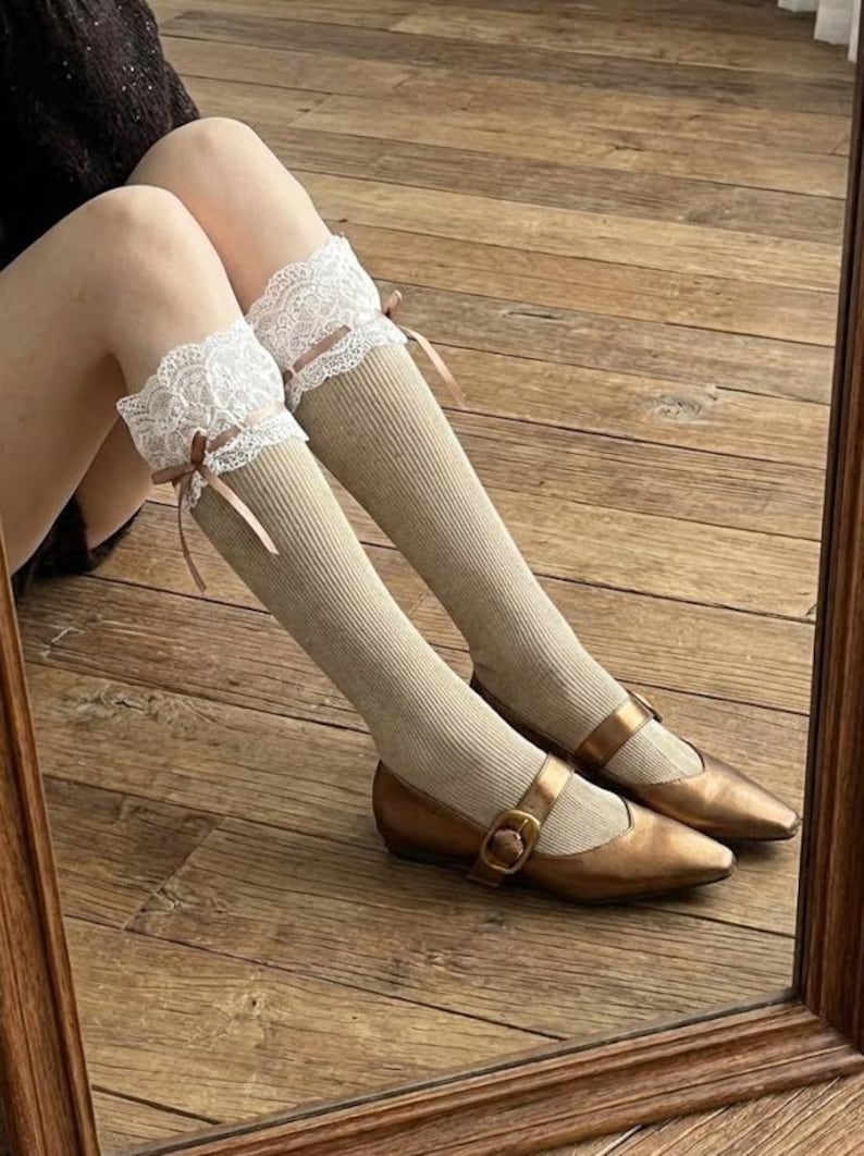 Pu&ograve; includere: Calzini al ginocchio a coste beige con finiture in pizzo bianco e piccoli fiocchi marroni. I calzini sono abbinati a scarpe Mary Jane color bronzo con chiusure con fibbia. L'immagine &egrave; ripresa da una prospettiva bassa, mostrando gambe e piedi.