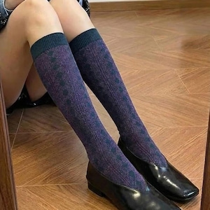 Puede incluir: Bailarinas de cuero negro con punta cuadrada, combinadas con calcetines hasta la rodilla. Los calcetines son morados con puño verde oscuro y un estampado de rombos. Los zapatos son brillantes y reflejan la luz.