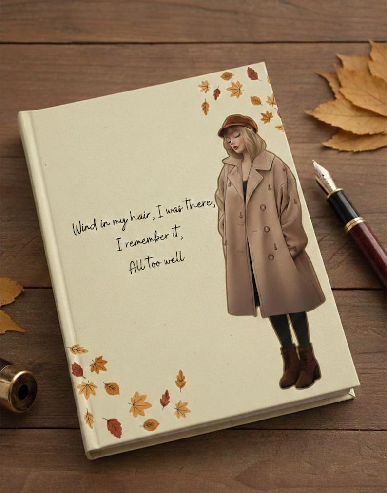May include: A cream-colored journal with the text "Wind in my hair, I was there, I remember it, All too well." features an illustration of a woman in a brown coat. Autumn leaves decorate the cover. A pen rests on the wooden surface.