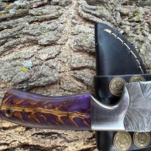 May include: A handmade knife with a purple and brown handle and a patterned steel blade. The knife is partially sheathed in a black leather holder with decorative metal accents. The handle features a unique wood grain design.