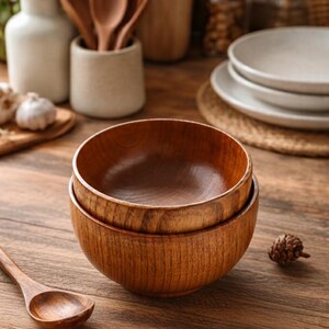 Puede incluir: Dos cuencos de madera apilados, mostrando un color marr&oacute;n c&aacute;lido. Los cuencos tienen un interior liso y un exterior texturizado. Una cuchara de madera descansa cerca, junto con otros utensilios de cocina, sugiriendo una est&eacute;tica r&uacute;stica y natural.