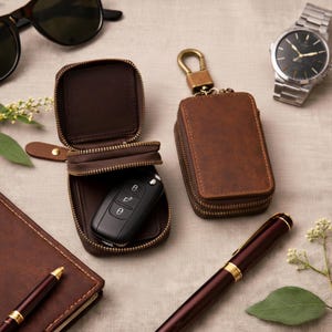 May include: A brown leather key case with a zip, open to show a car key. A closed key case with a brass clasp is next to it. Other accessories include a watch, sunglasses, a pen, and a notebook.