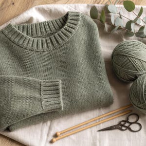 Può includere: Un maglione a maniche lunghe color verde salvia con girocollo a coste e polsini a coste. Due gomitoli di lana abbinati, un paio di ferri da maglia in legno e forbici in metallo in stile vintage sono disposti accanto al capo in maglia piegato.