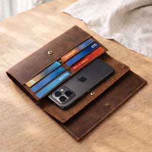 May include: Open brown leather wallet with card slots holding credit cards from Citi, UnioBank, and Wells Fargo. A black smartphone with the Apple logo is inside. The wallet is open on a wooden surface.