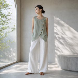 May include: A sage green sleeveless top featuring an asymmetrical overlapping front panel secured with a row of light brown buttons, paired with loose-fitting white wide-leg trousers and simple tan flat sandals for a coordinated outfit.