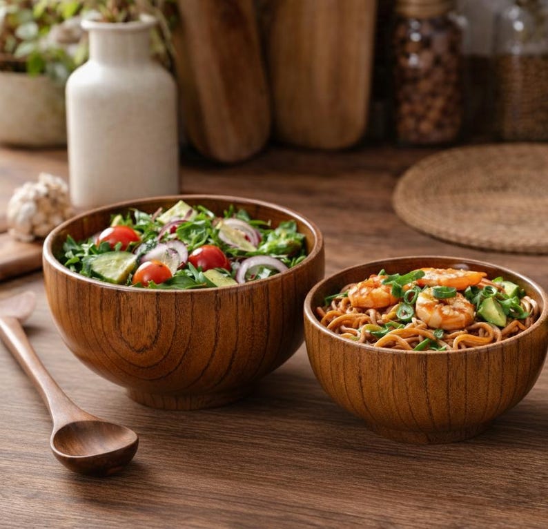 Puede incluir: Dos cuencos de madera llenos de comida sobre una mesa de madera. Uno contiene una ensalada con tomates, pepinos y cebolla roja. El otro tiene fideos con camarones y cebolletas. Una cuchara de madera descansa sobre la mesa.