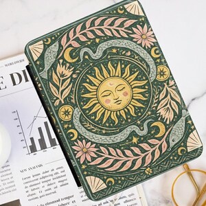 Könnte beinhalten: Eine grüne Tablet-Hülle mit einem Sonnenmotiv, umgeben von Schlangen, Blumen und Monden in Gelb, Rosa und Hellgrün. Die Hülle liegt auf einer Marmoroberfläche neben einer Zeitung und einer Kaffeetasse.