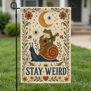 May include: A rectangular garden flag featuring a green frog playing a guitar while riding a blue snail. The design includes a crescent moon, stars, an eye, and floral borders in orange and green. The text STAY WEIRD is printed in dark blue.