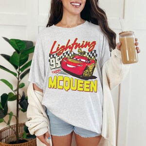 Puede incluir: Camiseta gris claro con un gr&aacute;fico de Rayo McQueen de la pel&iacute;cula Cars. El dise&ntilde;o incluye el texto "Lightning McQueen 95 Cars" en rojo y amarillo. La camiseta se combina con pantalones cortos de mezclilla y una rebeca color crema.