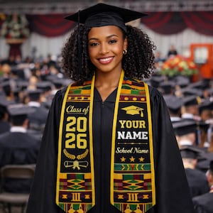 May include: A graduate in a black cap and gown smiles at the camera, wearing a graduation stole. The stole is black with gold trim and has the text "Class of 2026" and space for a name and school. A graduation ceremony is in the background.