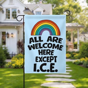 May include: A light blue garden flag with a rainbow graphic and the text "ALL ARE WELCOME HERE EXCEPT I.C.E." The flag is hanging on a black metal stand in a grassy yard, with a house in the background.