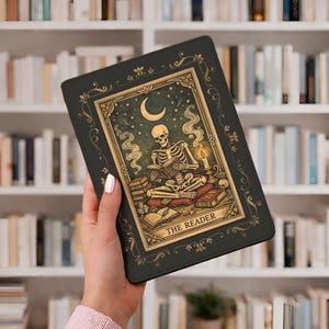 Peut inclure: Une housse de tablette noire avec un motif de carte de tarot. La carte représente un squelette lisant un livre sous un croissant de lune et des étoiles, encadré de détails dorés. Le texte "THE READER" est en bas de la carte.