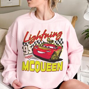 Puede incluir: Una sudadera rosa claro con un dise&ntilde;o de Rayo McQueen de la pel&iacute;cula Cars. El dise&ntilde;o incluye el texto "Lightning McQueen", "95" y "Cars" en rojo, amarillo y negro. La sudadera es de cuello redondo.