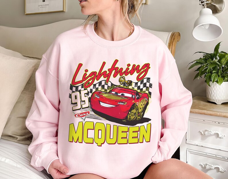 Puede incluir: Una sudadera rosa claro con un dise&ntilde;o de Rayo McQueen de la pel&iacute;cula Cars. El dise&ntilde;o incluye el texto "Lightning McQueen", "95" y "Cars" en rojo, amarillo y negro. La sudadera es de cuello redondo.