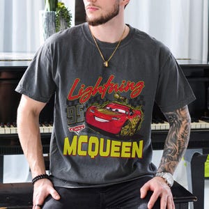 Puede incluir: Camiseta gris oscuro con un dise&ntilde;o de Rayo McQueen de la pel&iacute;cula Cars. El dise&ntilde;o incluye el texto "Lightning McQueen" en rojo y amarillo, con el n&uacute;mero "95" y la palabra "Cars".