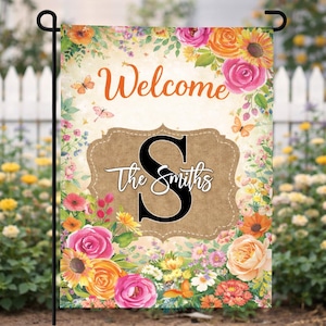 May include: A garden flag with a floral design and the word "Welcome" in orange script. The flag features a burlap-style emblem with a black letter "S" and the name "The Smiths" in white. Butterflies and flowers in shades of pink, orange, and yellow surround the text.