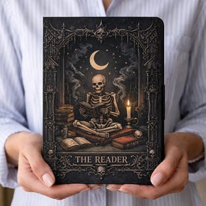 May include: A black tablet case featuring a gothic illustration of a skeleton reading a book under a crescent moon. The design includes a lit candle, skulls, and the text "THE READER". The case is held in front of a light blue shirt.
