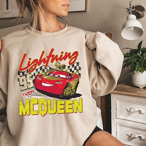 Puede incluir: Sudadera beige con un gr&aacute;fico de Rayo McQueen de la pel&iacute;cula Cars. El dise&ntilde;o incluye el texto "Lightning McQueen 95 Cars" en rojo y amarillo. La sudadera tiene mangas largas y cuello redondo.